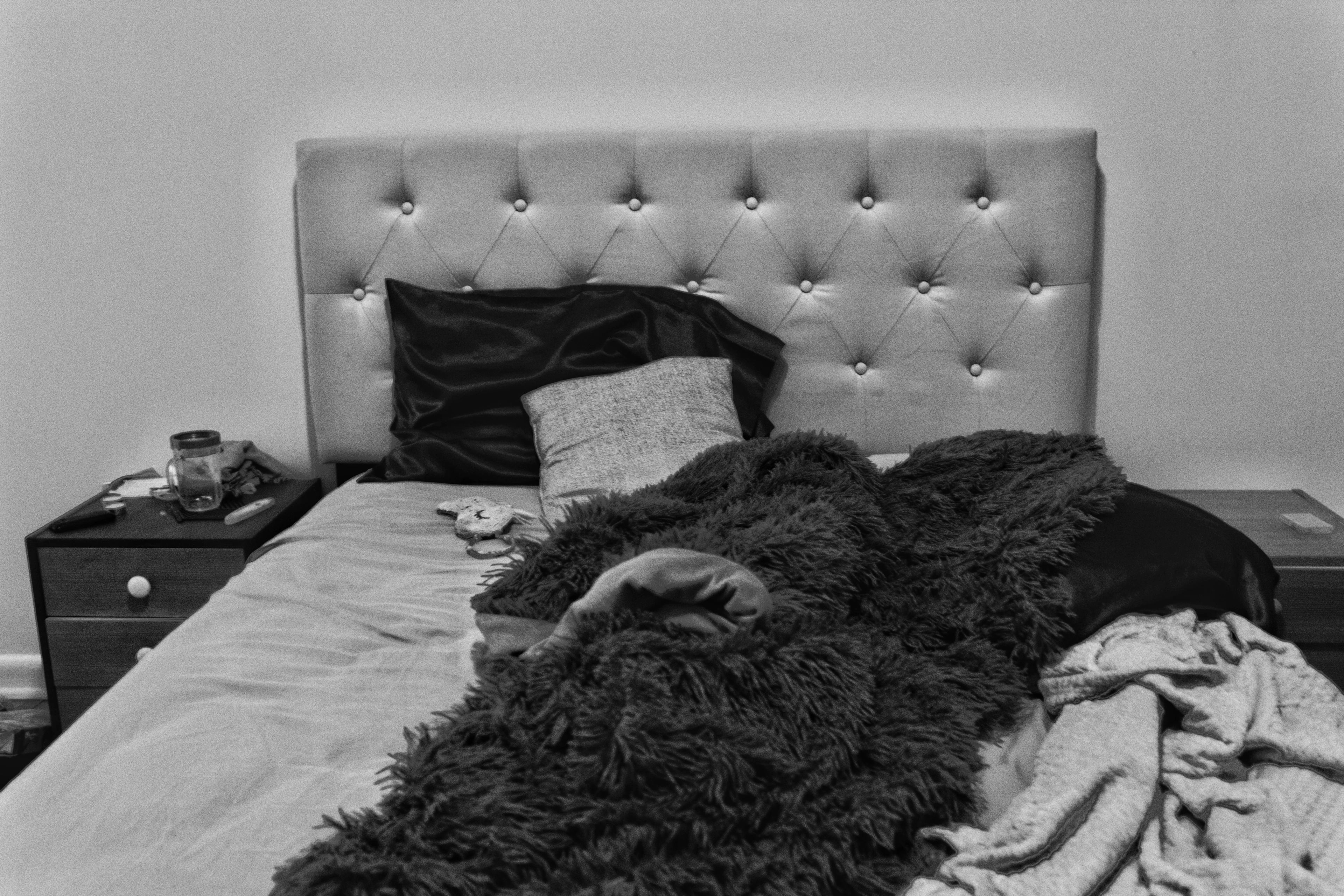 Black-and-white photograph — an unmade bed with a tufted headboard and a dark throw