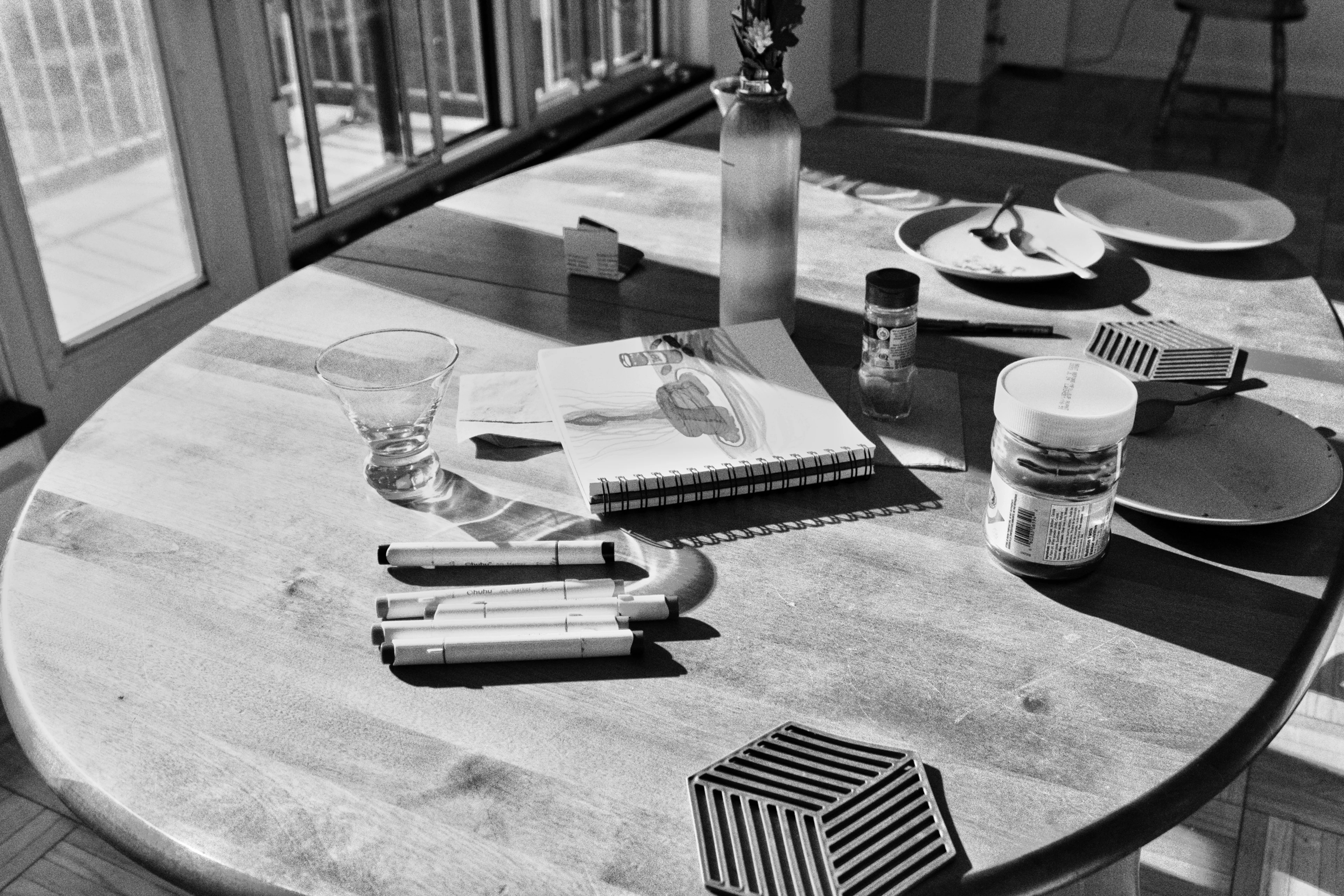 Black-and-white photograph — a round wooden table with notebooks, a glass and a small striped box