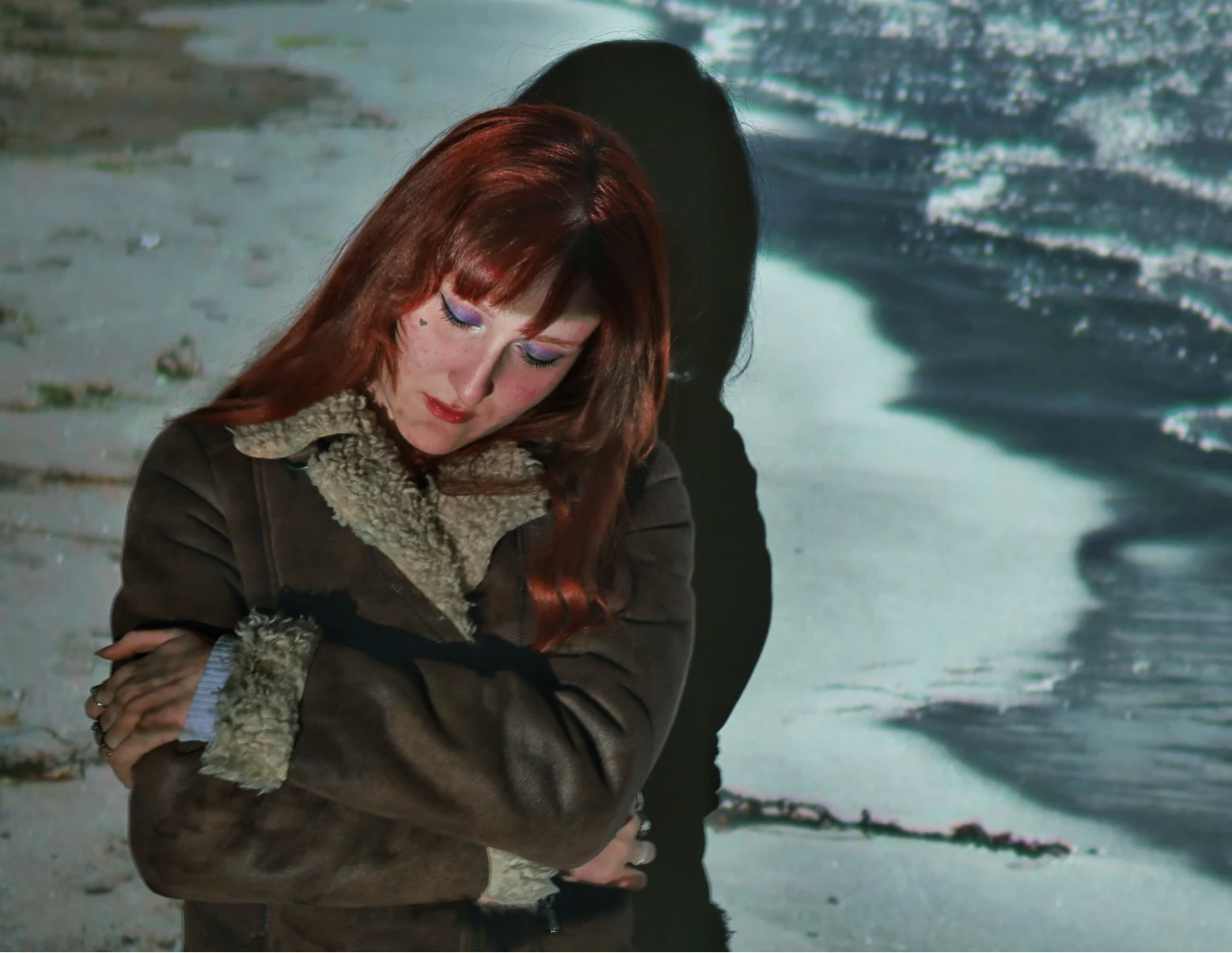 Photograph — a woman in a shearling jacket sitting on a beach, hugging her knees