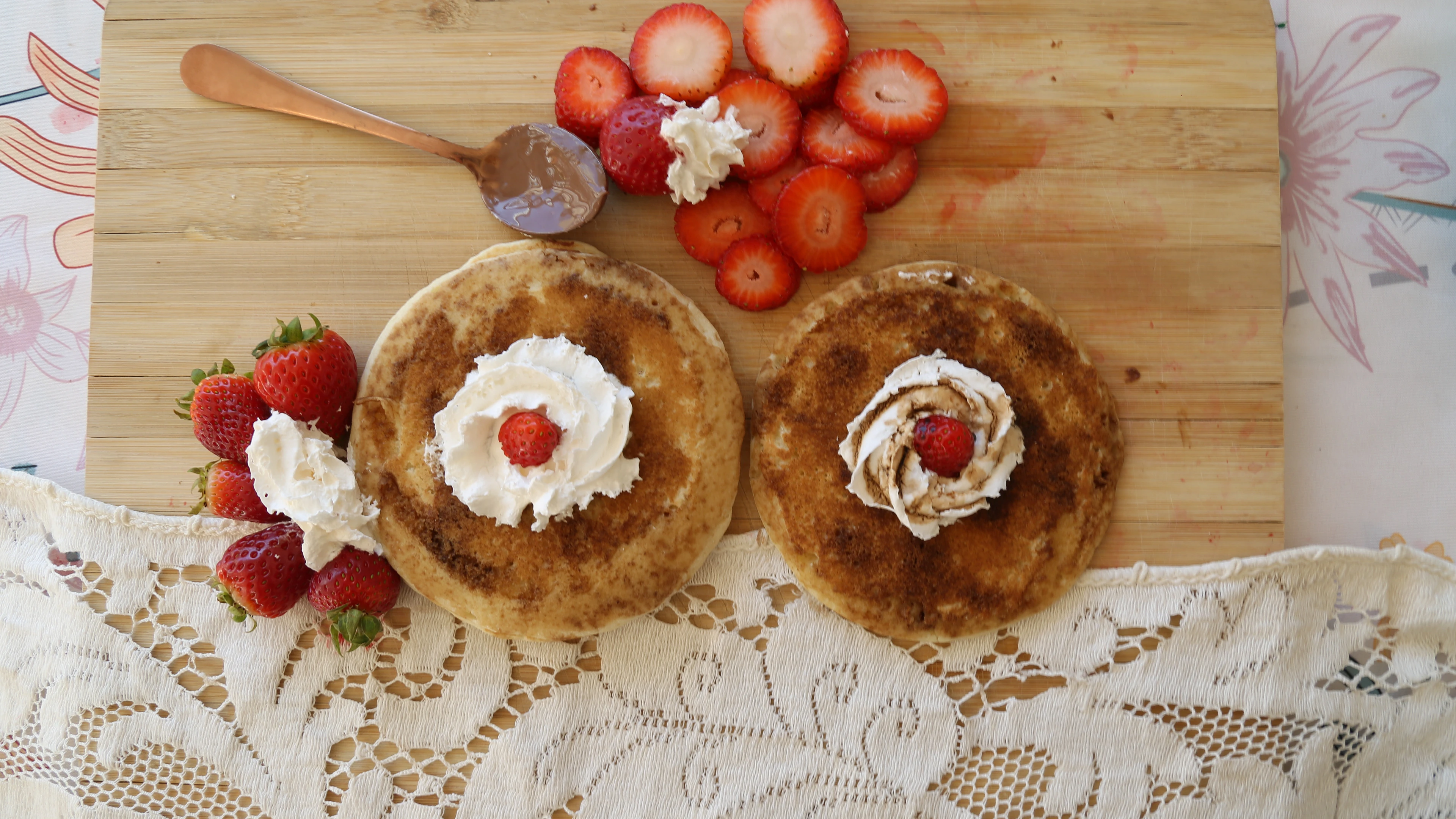 Food photograph — two pancakes topped with cream and strawberries on a board over a lace cloth