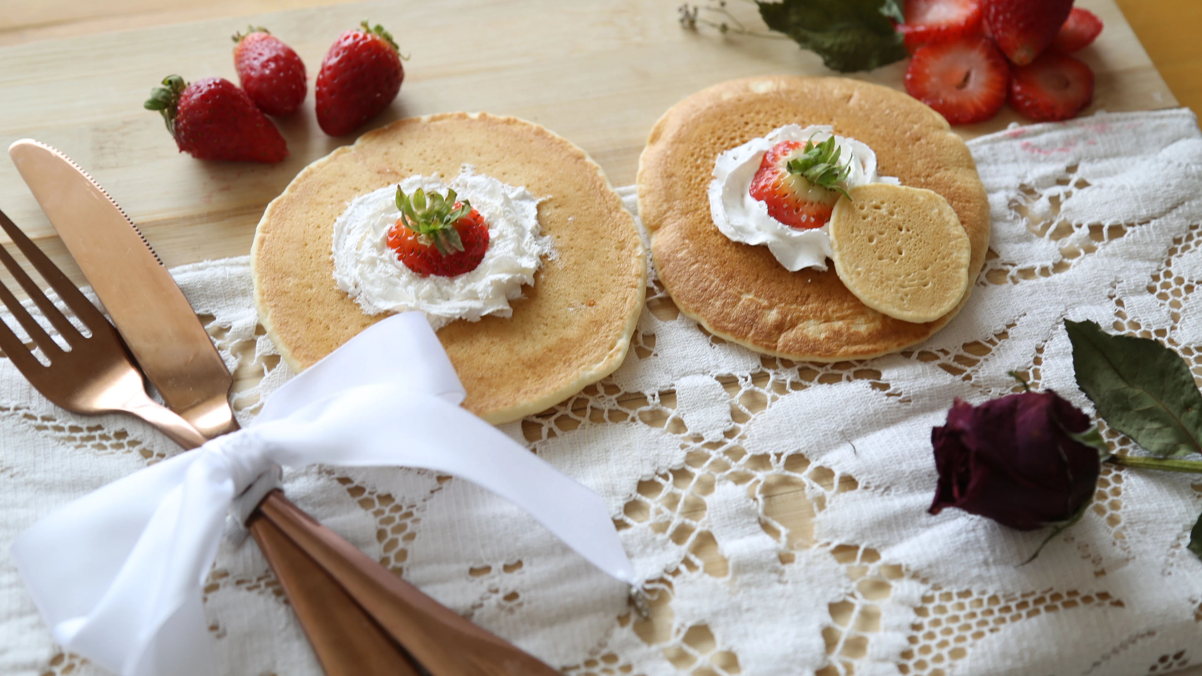 Food photograph — pancakes with cream and strawberries, copper cutlery tied with a white ribbon and a dark red rose