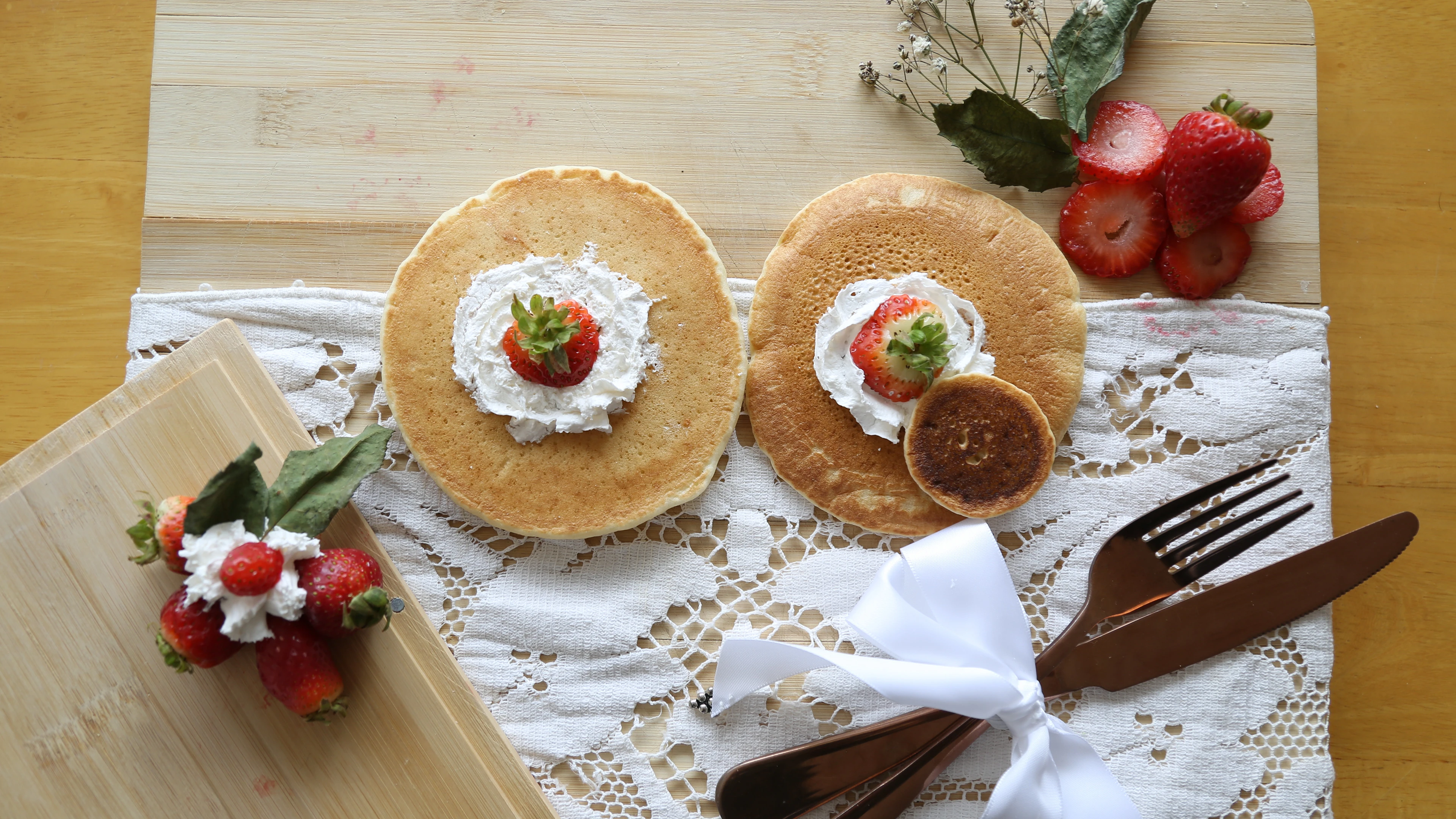 Food photograph — pancakes with cream and strawberries, a tray of berries, ribboned cutlery and a small flower arrangement