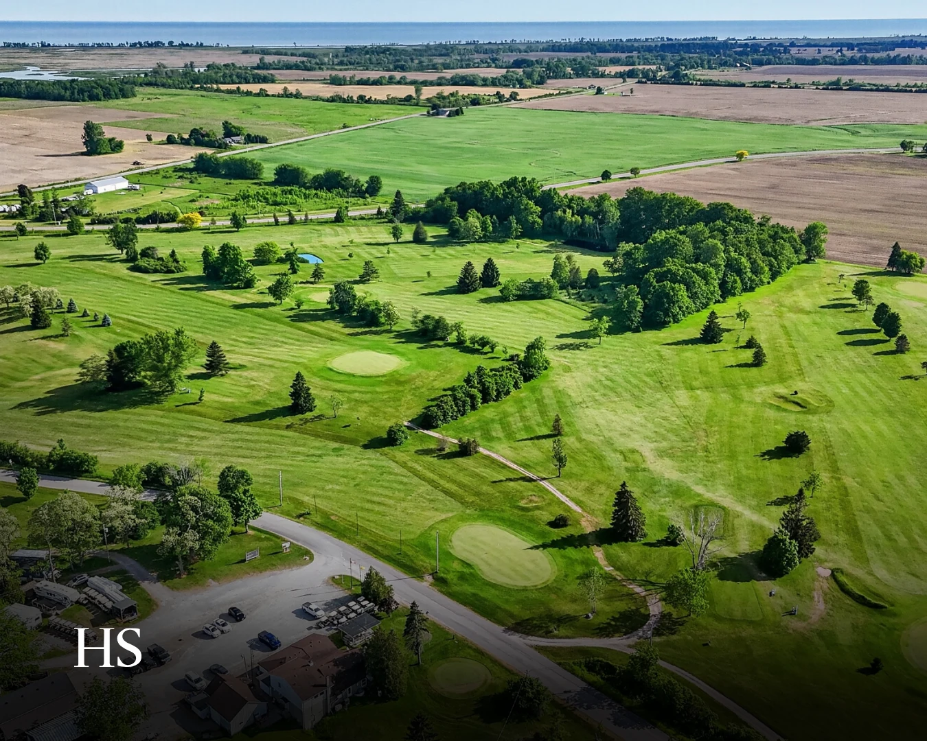 Drone aerial over the farmland and tree line beyond the neighbourhood, water at the horizon