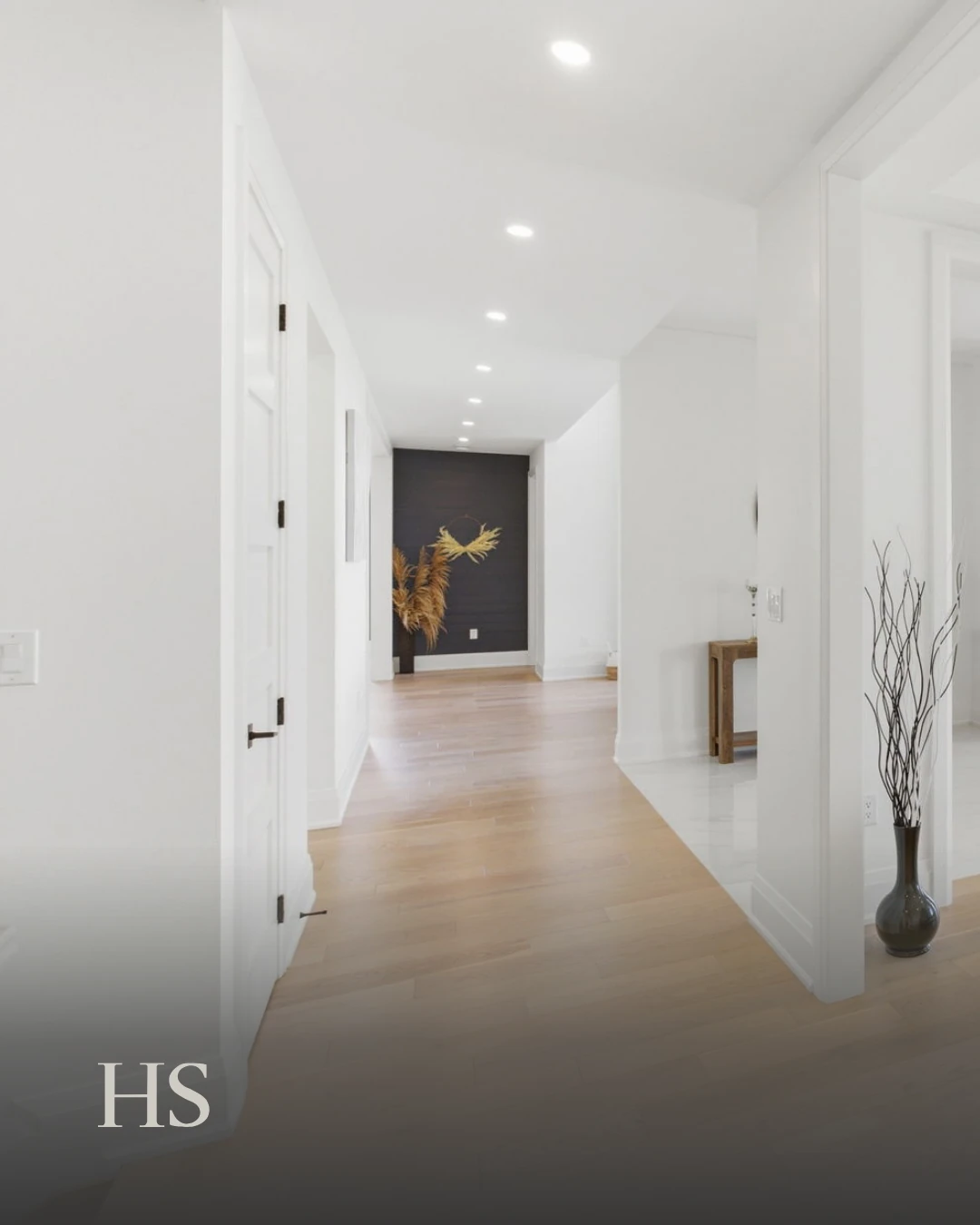 White hallway with a wood floor leading to a dark fireplace and a vase of bare branches