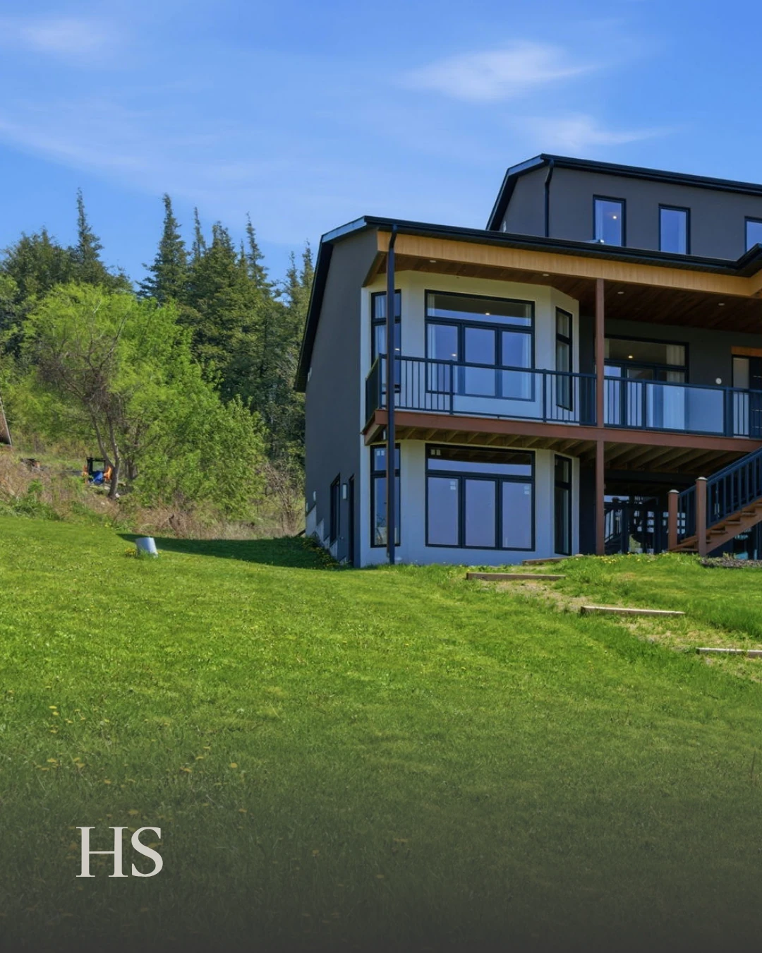 The charcoal multi-level home on a sloping green lawn, with wood soffit, balconies and evergreens