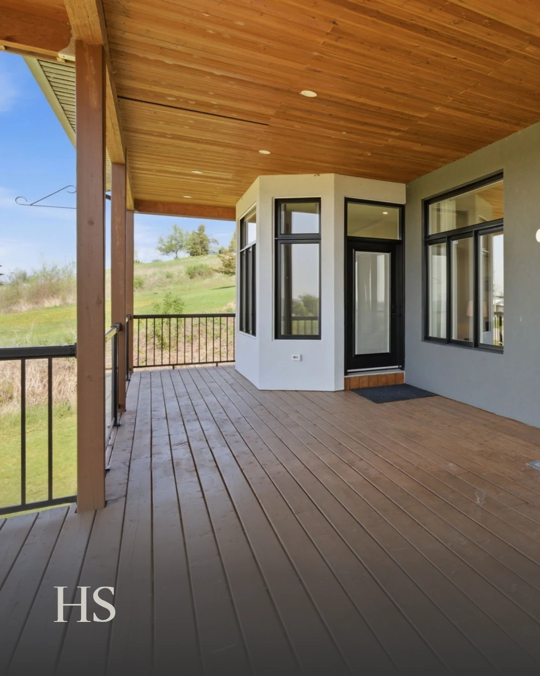 Covered deck with a wood-plank ceiling and recessed lights, a black railing and a dark-framed glass door, overlooking a grassy hillside